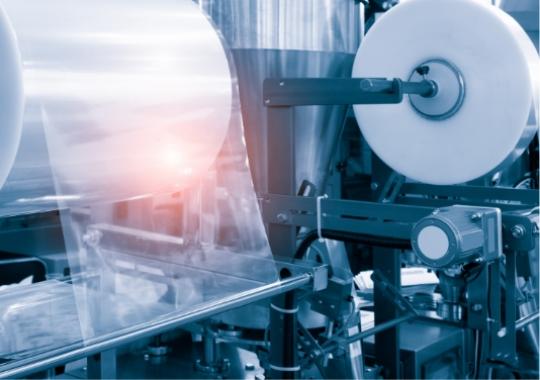 Macchinario industriale avvolge plastica trasparente, riflessi metallici e atmosfera tecnologica futuristica.